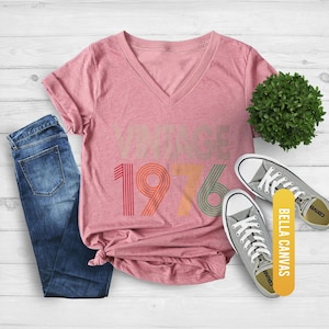 May include: A heather mauve V-neck t-shirt with the text "VINTAGE 1976" in retro colors. The shirt is styled with blue jeans and gray sneakers. A small green plant and a "BELLA CANVAS" tag are also present.
