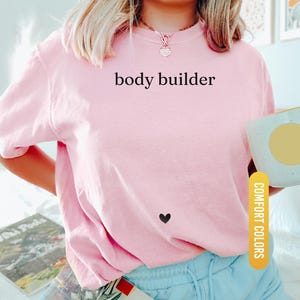 Pu&ograve; includere: Una maglietta rosa chiaro con le parole "body builder" stampate in nero. Un piccolo cuore nero &egrave; sulla maglietta. La maglietta fa parte della collezione Comfort Colors.