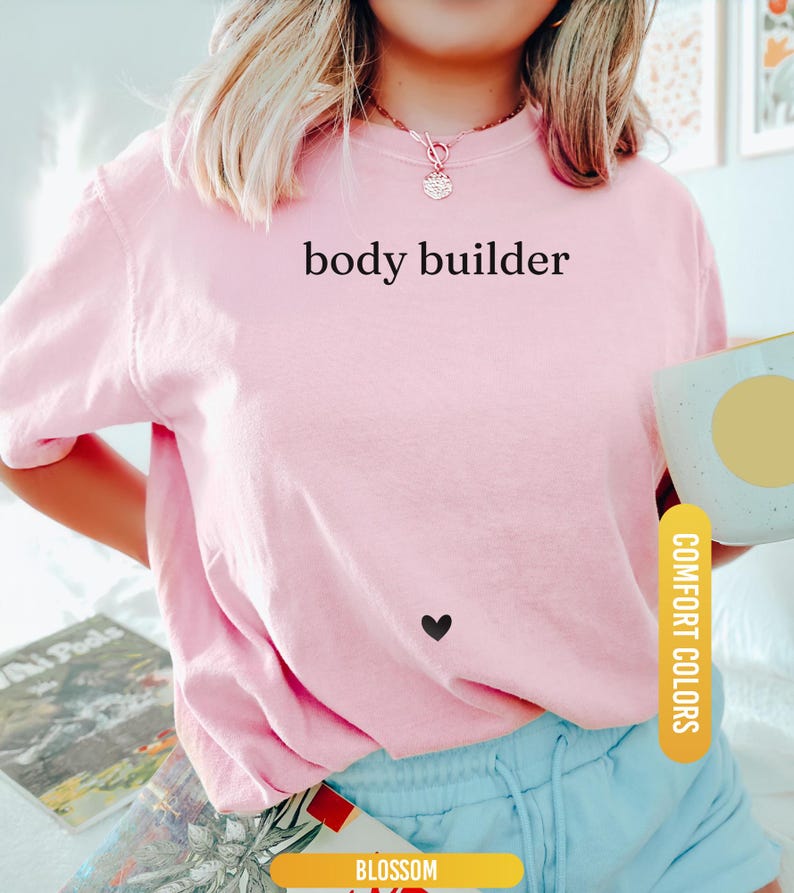 Pu&ograve; includere: Una maglietta rosa chiaro con le parole "body builder" stampate in nero. Un piccolo cuore nero &egrave; sulla maglietta. La maglietta fa parte della collezione Comfort Colors.