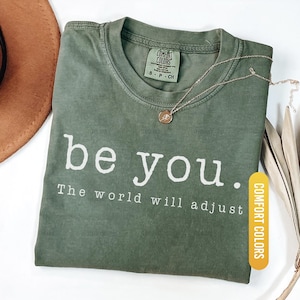 May include: A moss green t-shirt with the text "be you. The world will adjust" in white. The shirt has a crew neck and short sleeves. A gold necklace with a small pendant rests on the shirt. The shirt is labeled "Comfort Colors".