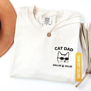 May include: Ivory-colored t-shirt with the text "CAT DAD" and a cat wearing sunglasses. The shirt also features the names "Millie" and "Ollie" below the cat graphic. A gold necklace and a brown hat are also visible.