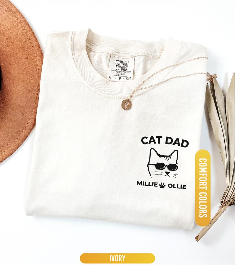 May include: Ivory-colored t-shirt with the text "CAT DAD" and a cat wearing sunglasses. The shirt also features the names "Millie" and "Ollie" below the cat graphic. A gold necklace and a brown hat are also visible.