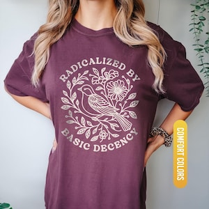 May include: Berry-colored t-shirt with a graphic that reads "RADICALIZED BY BASIC DECENCY" around a bird and floral design. The shirt is labeled "COMFORT COLORS" and "BERRY".
