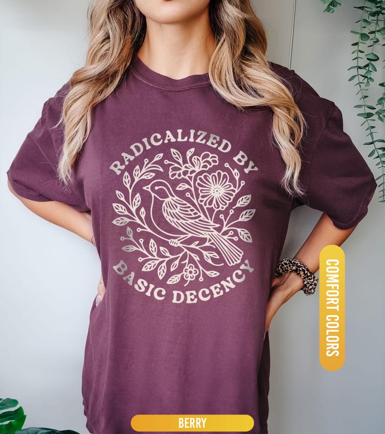 May include: Berry-colored t-shirt with a graphic that reads "RADICALIZED BY BASIC DECENCY" around a bird and floral design. The shirt is labeled "COMFORT COLORS" and "BERRY".