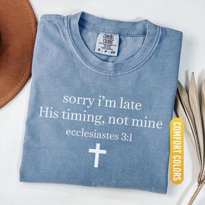 May include: Blue jean Comfort Colors t-shirt with the text "sorry i'm late His timing, not mine ecclesiastes 3:1" and a white cross. The shirt is folded and has a tag that says "Comfort Colors".