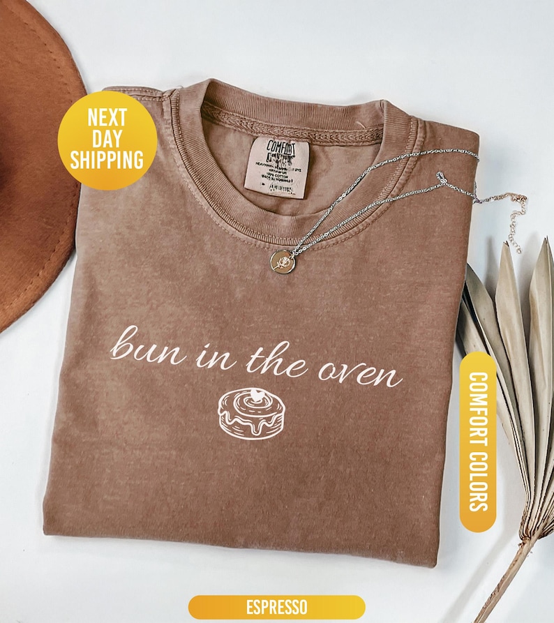 May include: Espresso-colored t-shirt with the phrase "bun in the oven" and a graphic of a cinnamon roll. A silver necklace with a pendant rests on the shirt. The shirt is folded and displayed with a hat and dried plant.
