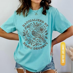 May include: A turquoise t-shirt with the text "RADICALIZED BY BASIC DECENCY" in brown, surrounding a bird and floral design. The shirt is a comfort color and is labeled "CHALKY MINT".
