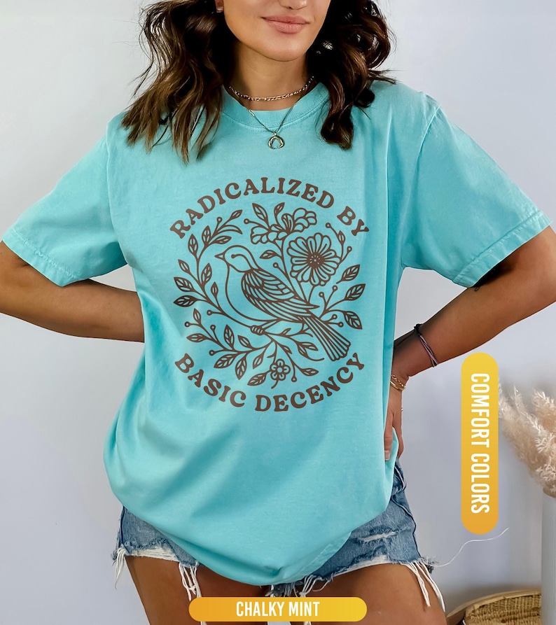 May include: A turquoise t-shirt with the text "RADICALIZED BY BASIC DECENCY" in brown, surrounding a bird and floral design. The shirt is a comfort color and is labeled "CHALKY MINT".