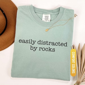 May include: A light green t-shirt with the text "easily distracted by rocks" in black. The shirt has a crew neck and a tag that says "Comfort Colors". A gold necklace and a brown hat are also in the image.