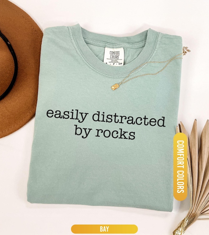 May include: A light green t-shirt with the text "easily distracted by rocks" in black. The shirt has a crew neck and a tag that says "Comfort Colors". A gold necklace and a brown hat are also in the image.