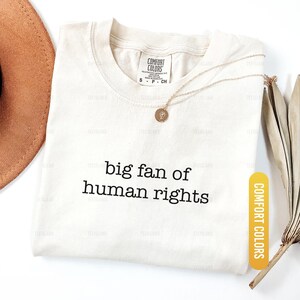 May include: Ivory-colored t-shirt with the text "big fan of human rights" in black. A gold necklace with a pendant rests on the shirt. The shirt has a Comfort Colors tag and an Ivory color label.