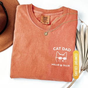May include: A yam-colored Comfort Colors t-shirt with the words "CAT DAD" and a cat graphic wearing sunglasses. The shirt also features the names "Millie" and "Ollie". A gold necklace and a brown hat are also visible.