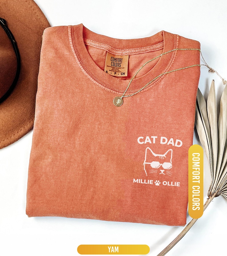 May include: A yam-colored Comfort Colors t-shirt with the words "CAT DAD" and a cat graphic wearing sunglasses. The shirt also features the names "Millie" and "Ollie". A gold necklace and a brown hat are also visible.
