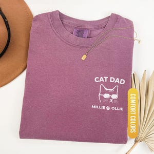 May include: Berry-colored t-shirt with the text "CAT DAD" and a cat wearing sunglasses. The shirt also features the names "Millie" and "Ollie" below the cat graphic. A gold necklace and a brown hat are also visible.