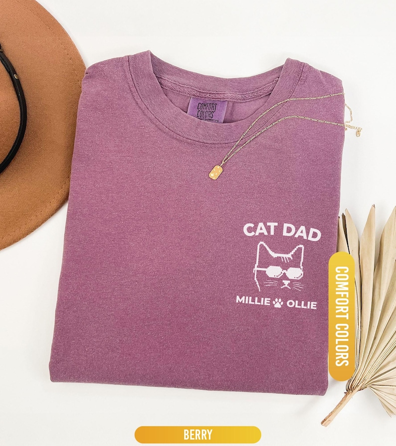 May include: Berry-colored t-shirt with the text "CAT DAD" and a cat wearing sunglasses. The shirt also features the names "Millie" and "Ollie" below the cat graphic. A gold necklace and a brown hat are also visible.