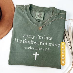May include: A moss green Comfort Colors t-shirt with the text "sorry I'm late His timing, not mine ecclesiastes 3:1" and a white cross. A gold necklace with a pendant is draped on the shirt.