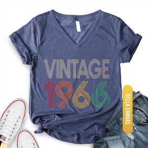 Pu&ograve; includere: T-shirt con scollo a V blu navy melange con la scritta "VINTAGE 1966" in un design retr&ograve;. I numeri sono rossi, arancioni e verdi. La maglietta &egrave; abbinata a scarpe da ginnastica bianche e jeans blu.