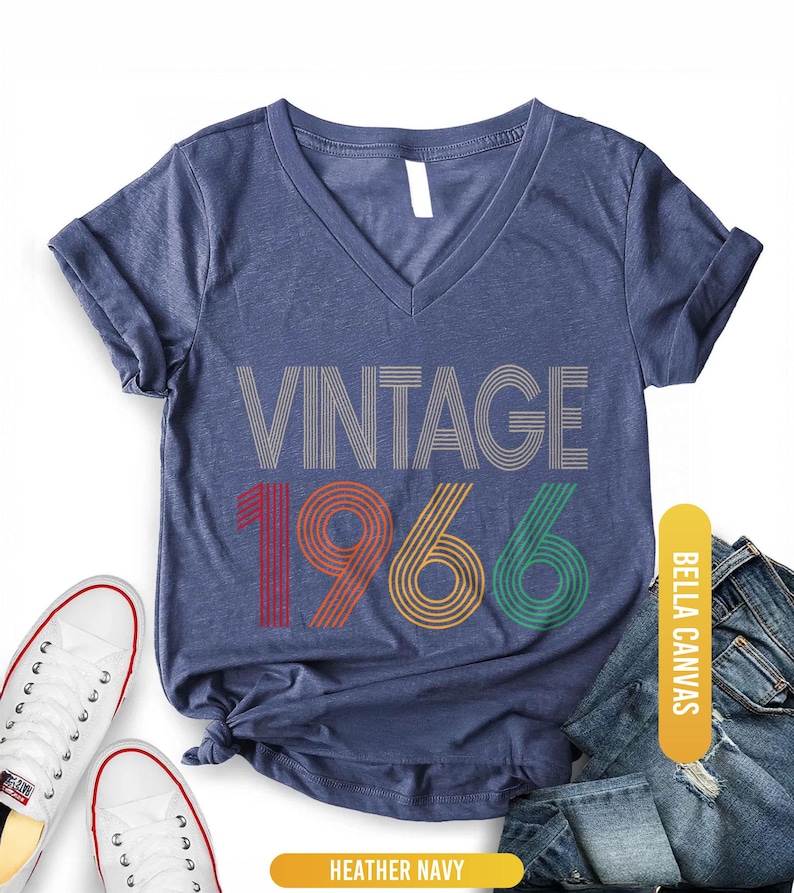 Pu&ograve; includere: T-shirt con scollo a V blu navy melange con la scritta "VINTAGE 1966" in un design retr&ograve;. I numeri sono rossi, arancioni e verdi. La maglietta &egrave; abbinata a scarpe da ginnastica bianche e jeans blu.