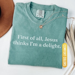 May include: Light green Comfort Colors t-shirt with the text "First of all, Jesus thinks I'm a delight." A gold-colored necklace with a pendant rests on the shirt. The shirt is labeled "Light Green."
