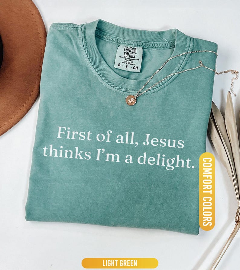 May include: Light green Comfort Colors t-shirt with the text "First of all, Jesus thinks I'm a delight." A gold-colored necklace with a pendant rests on the shirt. The shirt is labeled "Light Green."