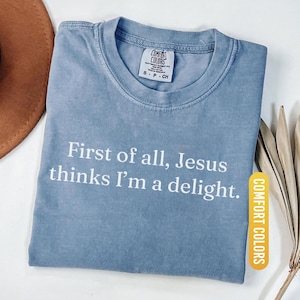 May include: Blue jean Comfort Colors t-shirt with the text "First of all, Jesus thinks I'm a delight." The shirt has a crew neck and short sleeves. The Comfort Colors label is visible on the upper chest.