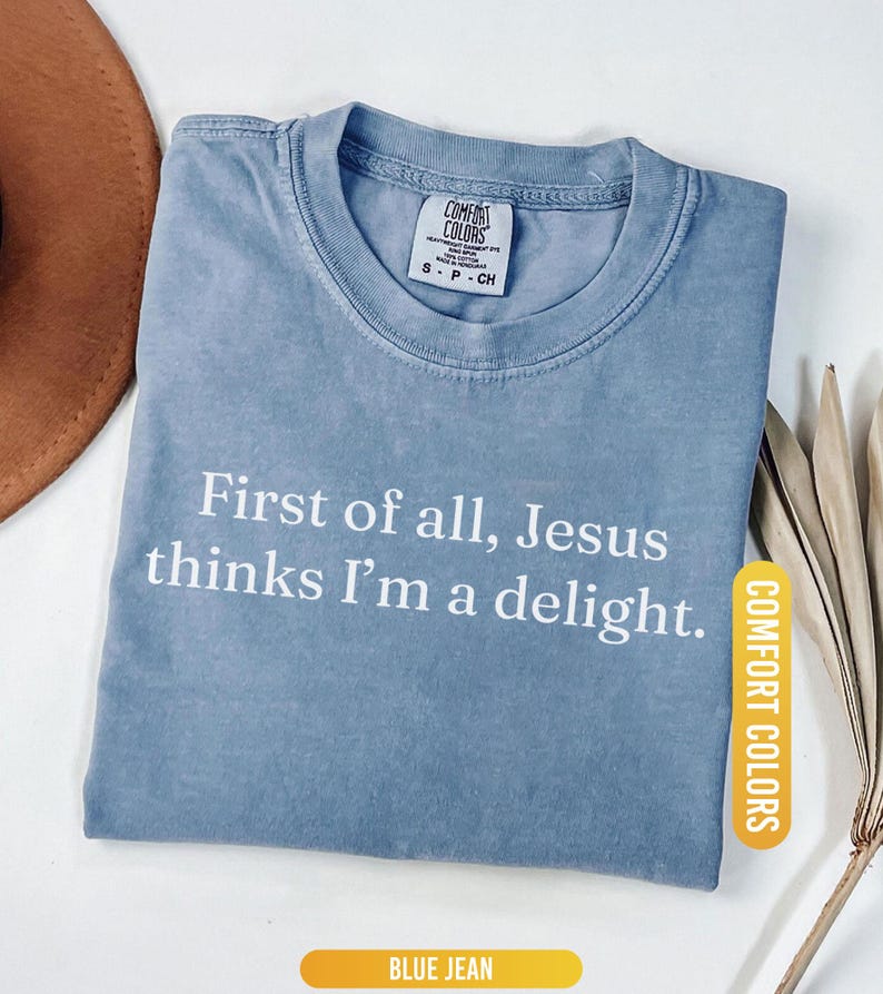 May include: Blue jean Comfort Colors t-shirt with the text "First of all, Jesus thinks I'm a delight." The shirt has a crew neck and short sleeves. The Comfort Colors label is visible on the upper chest.