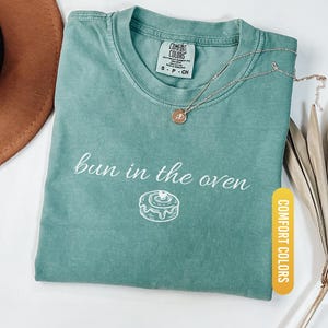 May include: Light green Comfort Colors t-shirt with the phrase "bun in the oven" and a graphic of a cinnamon roll. A gold necklace with a pendant rests on the shirt. The shirt is folded and displayed with a brown hat and dried plants.