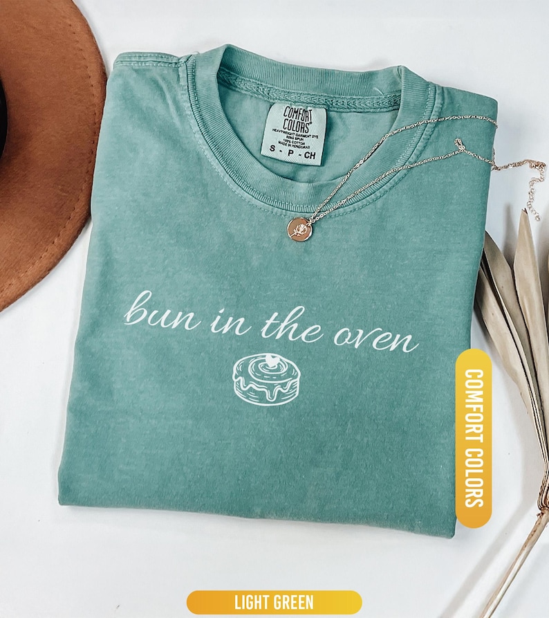May include: Light green Comfort Colors t-shirt with the phrase "bun in the oven" and a graphic of a cinnamon roll. A gold necklace with a pendant rests on the shirt. The shirt is folded and displayed with a brown hat and dried plants.