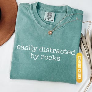 May include: Light green Comfort Colors t-shirt with the text "easily distracted by rocks" in white. A gold-colored necklace with a pendant rests on the shirt. The shirt is folded and has a tag.