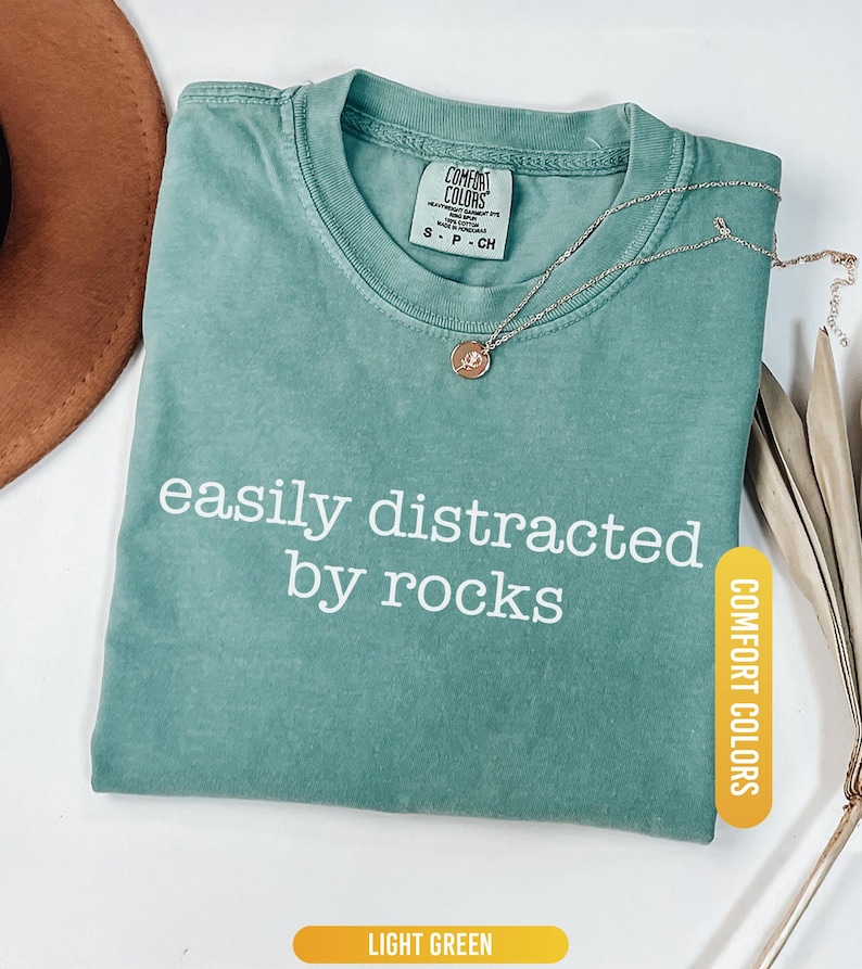 May include: Light green Comfort Colors t-shirt with the text "easily distracted by rocks" in white. A gold-colored necklace with a pendant rests on the shirt. The shirt is folded and has a tag.