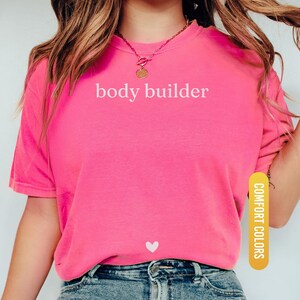 Pu&ograve; includere: Una maglietta rosa acceso con la scritta bianca "body builder". Un piccolo cuore bianco &egrave; sotto il testo. La maglietta &egrave; abbinata a jeans blu. Le parole "Comfort Colors" e "Crunchberry" sono stampate sulla maglietta.