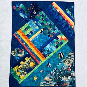 May include: A colorful quilt with a blue background featuring a variety of fish and coral designs. The quilt is made up of different fabric squares and strips, creating a patchwork effect.