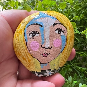 May include: Hand-painted rock art featuring a stylized face with blonde hair, blue accents, and pink cheeks. The rock is held in a hand, with green foliage and a wooden surface in the background. The artwork has a whimsical, folk art style.
