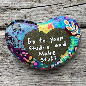 May include: A heart-shaped painted rock with a colorful abstract design. The rock has a black heart shape painted on it with the text "Go to your studio and Make Stuff" written in white. The text "Fred Babb" is written in white on the bottom right corner of the rock.