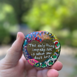 Puede incluir: Una piedra pintada con forma de coraz&oacute;n con el texto "Lo &uacute;nico imposible es lo que no intentas". La piedra est&aacute; decorada con puntos y remolinos de colores.