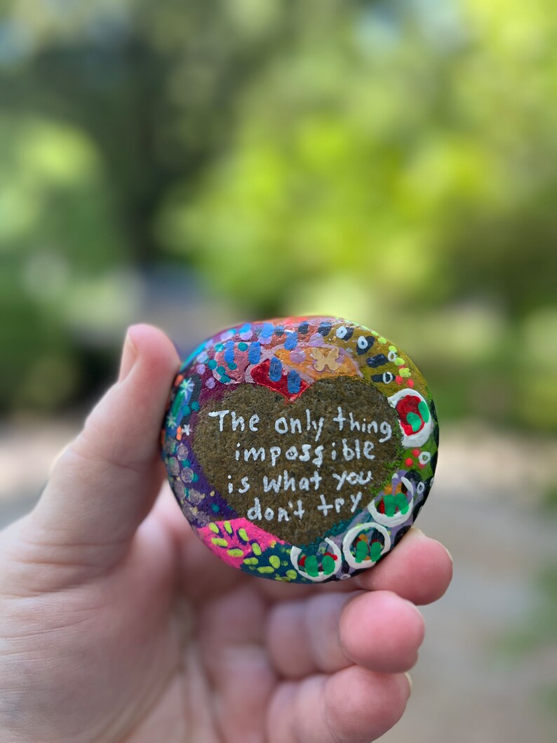 Puede incluir: Una piedra pintada con forma de coraz&oacute;n con el texto "Lo &uacute;nico imposible es lo que no intentas". La piedra est&aacute; decorada con puntos y remolinos de colores.