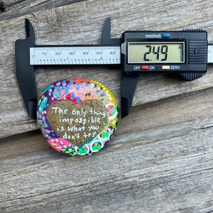 Puede incluir: Una piedra pintada de colores vivos con la cita "Lo &uacute;nico imposible es lo que no intentas" escrita en ella. La piedra se est&aacute; midiendo con un calibre, mostrando una medida de 6,3 cm.