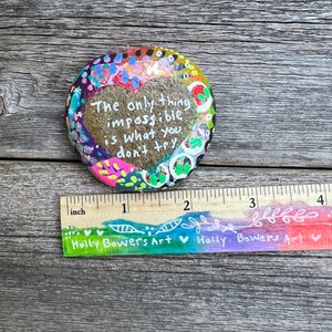 Puede incluir: Una piedra pintada con un dise&ntilde;o en forma de coraz&oacute;n y el texto "The only thing impossible is what you don't try". La piedra tiene un di&aacute;metro de aproximadamente 5 cm y est&aacute; decorada con puntos y remolinos coloridos.