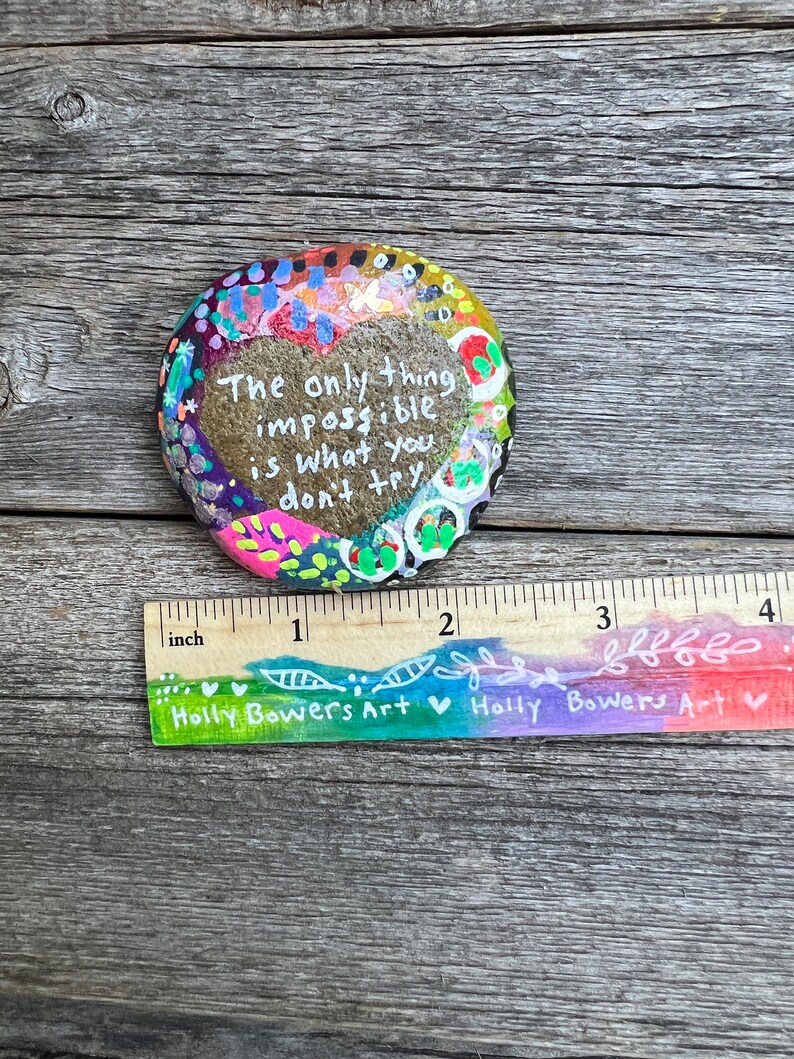 Puede incluir: Una piedra pintada con un dise&ntilde;o en forma de coraz&oacute;n y el texto "The only thing impossible is what you don't try". La piedra tiene un di&aacute;metro de aproximadamente 5 cm y est&aacute; decorada con puntos y remolinos coloridos.