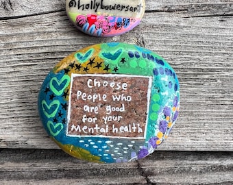 May include: Two painted rocks, one with the text "Choose people who are good for your mental health" and the other with the text "@hollybowersart".