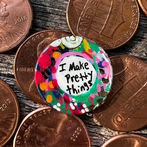 May include: A colorful, hand-painted button with the text "I Make Pretty Things" in black. The button is surrounded by a vibrant mix of red, pink, green, and yellow paint. It rests on a bed of U.S. pennies.
