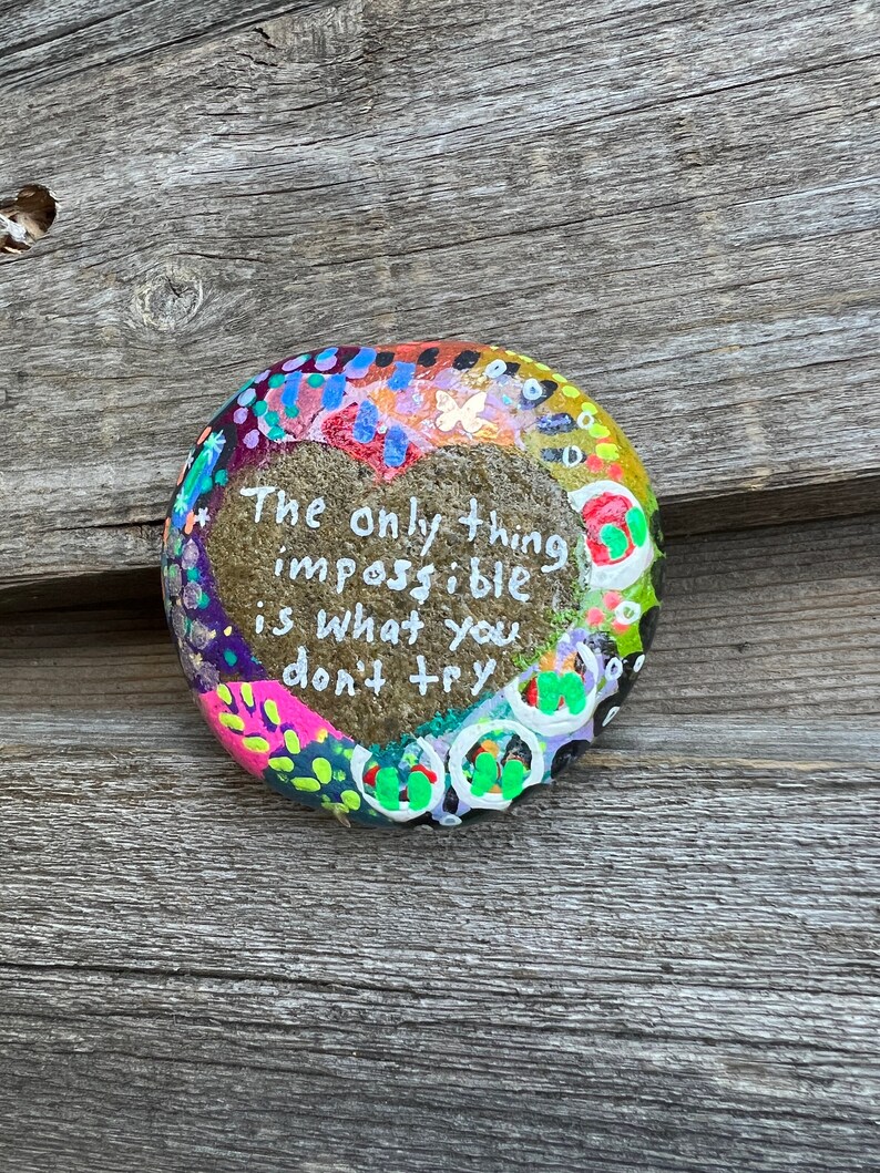 Puede incluir: Una piedra pintada con una forma de coraz&oacute;n en el centro. La piedra tiene un dise&ntilde;o colorido con puntos, remolinos y mariposas. El texto en la piedra dice "Lo &uacute;nico imposible es lo que no intentas."