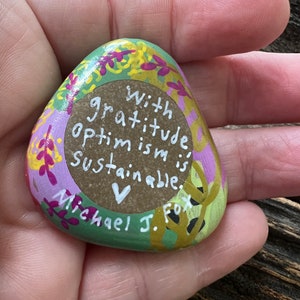 May include: A green, pink, and yellow painted rock with the words "With gratitude, optimism is sustainable." and a heart symbol. The rock is signed "Michael J. Fox."