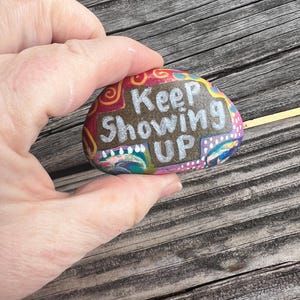 May include: A painted rock with the words "Keep Showing Up" in white paint on a brown background. The rock is decorated with colorful swirls and dots.