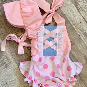 May include: Pink and blue baby outfit with a white lace trim and pink polka dots. The outfit includes a matching pink bonnet.
