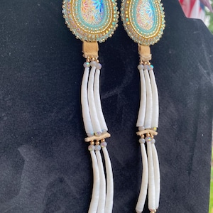 May include: A pair of long dangle earrings featuring teardrop-shaped components with iridescent centers, surrounded by gold and light blue beadwork. Below the teardrops are tan leather tabs, small grey beads, and long white tubular beads.