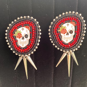 May include: A pair of circular earrings featuring a central white skull with a red rose and green leaves. The skull is surrounded by concentric rings of red and black beads, finished with a silver beaded edge and three dangling silver spikes.