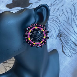 May include: A pair of black beaded earrings with pink and yellow accents. The earrings are round and have a black center with a starburst design.