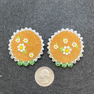 May include: A pair of round, brown suede earrings with white and yellow beaded flowers and green beaded leaves. The earrings are surrounded by white and green beads.
