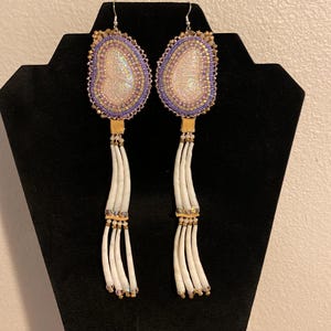 May include: Handmade beaded earrings with a light pink and purple oval design. The earrings have a white fringe and gold accents. The earrings are displayed on a black stand.